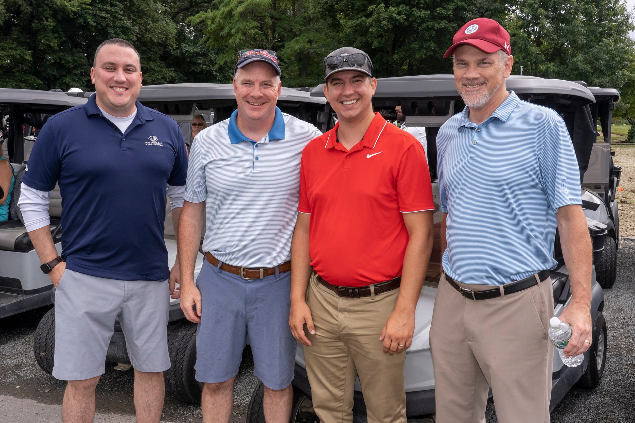 Four men stand oby golf carts, ready to go play golf. 